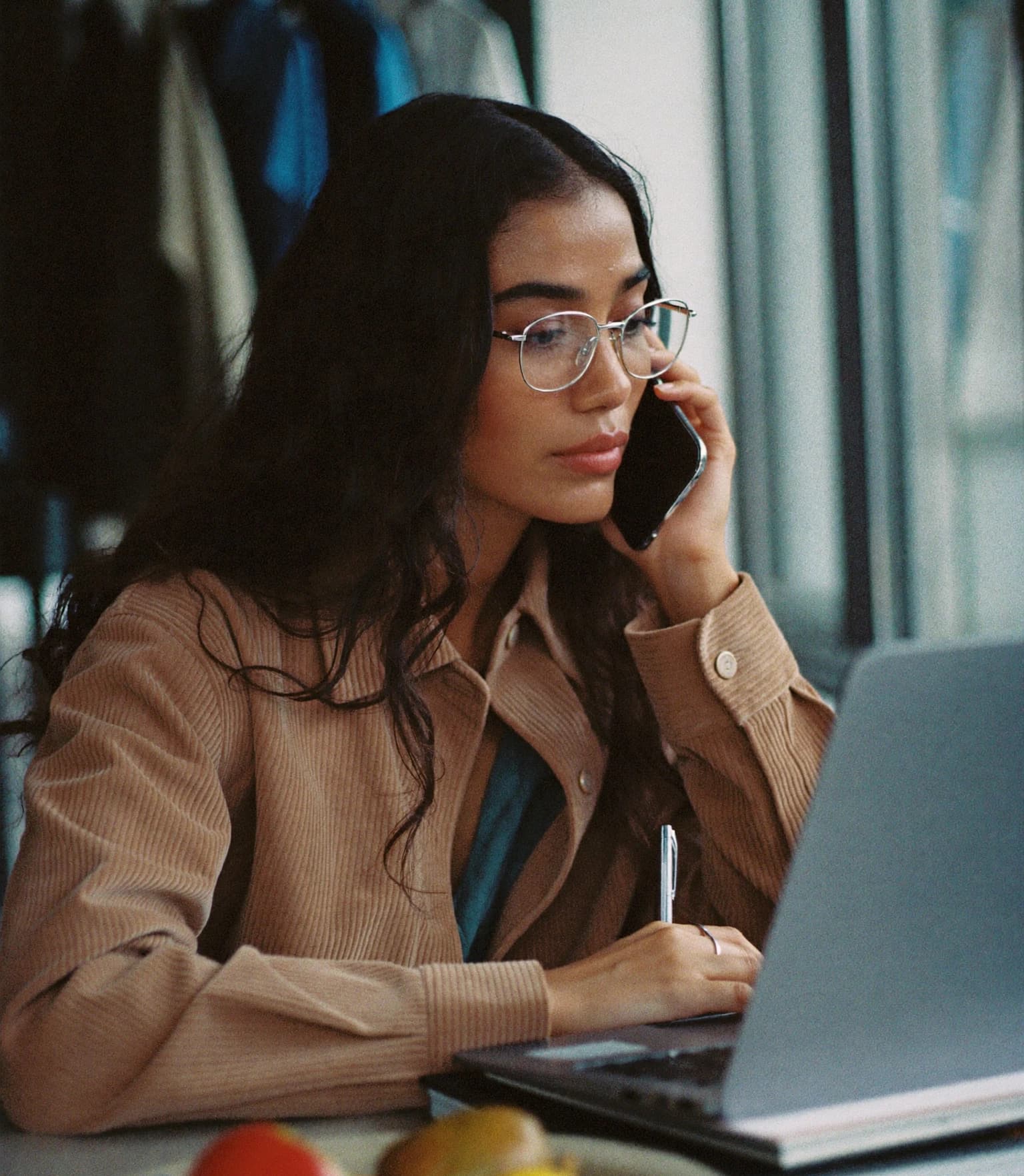 A fashion merchandiser on the phone at her laptop in a showroom, rails of clothing behind her.